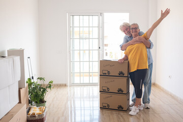 Wall Mural - Cheerful senior couple enjoys moving into a new home, surrounded by moving boxes in an empty room. The woman has outstretched arms, symbolizing excitement for their new beginning as retirees