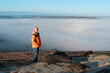 © Iryna - Man Standing on Rocky Peak Overlooking Sea of Fog on Sunny Winter Day