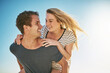 © Stratford/peopleimages.com - Happy couple, outdoor and piggyback for holiday, vacation and bonding with romance, fun and adventure. Excited young man and woman in Europe with support for tourism, travel and love on blue sky