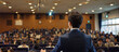© Lita Clarus - A man stands in front of a large audience, giving a speech