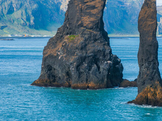 Aerial view of two large black rock formations creating a natural archway in the ocean with a hazy blue mountain range in the background, Iceland.