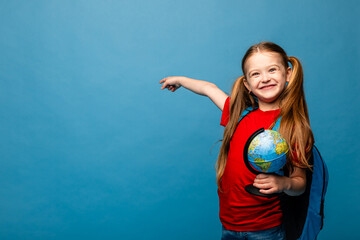 Wall Mural - Happy first year schoolgirl girl in a red t-shirt and jeans with a globe in her hands and backpack, point finger on empty blue background. Copy space. Back to school concept.