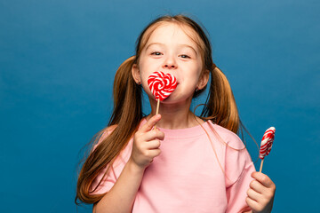 Wall Mural - Happiness, childhood, kid and sweets. A happy little girl holding a heart shaped lollipop caramel on a stick, A festive candy in the hands of child in pink t-shirt on blue background. Valentines Day