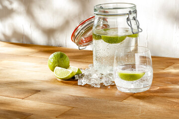 Wall Mural - Water with lime in a glass jug and a glass on a wooden table against a white wall and natural light. Advertising space