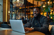 © Prasanth - Candid portrait of a black man working on a laptop in a stylish modern cafe hotel lobby, African american digital nomad on a virtual video call meeting with colleagues, generative ai