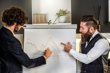 Businessmen analyzing financial chart on whiteboard. Two colleagues discussing data trends during a business meeting. Modern office environment.