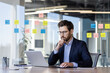 © Liubomir - Businessman working on laptop in modern office with sticky notes on glass wall. Focused professional in suit analyzing data, brainstorming ideas, and planning strategy for business success.