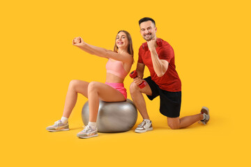 Wall Mural - Beautiful young happy couple in sportswear training with dumbbells and fitness ball on yellow background
