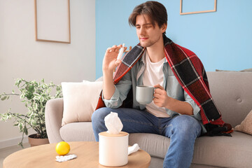 Sticker - Sick young man with nasal drops and cup of tea sitting on sofa at home