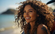 © imagineRbc - A close-up image of a woman with curly brown hair smiling at the beach. She is wearing a tank top and has a tattoo on her shoulder. The sun is shining and the water is visible in the background