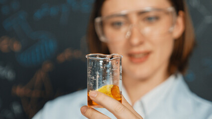 Poster - Closeup of young beautiful teacher holding chemical liquid at laboratory. Scientist doing an experiment by inspect colored solution while standing in front of blackboard. Focus on beaker. Erudition.