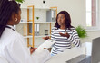 © Studio Romantic - Female african american doctor talking with a young worried sick woman patient and writing down her complaints sitting at the desk in office during medical examination in clinic. Health care concept.
