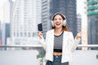 © Chanakon - Young asian woman using smartphone while walking in the urban city. Happy Beautiful asian female with headphone listen to the music by cell phone standing at office building city. Lifestyle people