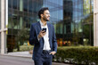© Liubomir - Confident young businessman in suit holding smartphone and smiling while standing outdoors near modern office building. Professional attitude, business success, and positive outlook convey success