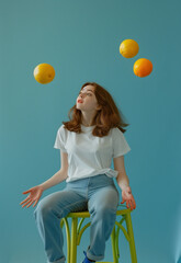 Wall Mural - A woman sitting on a blue chair, juggling balls and a peach in the air.Minimal creative fun concept.