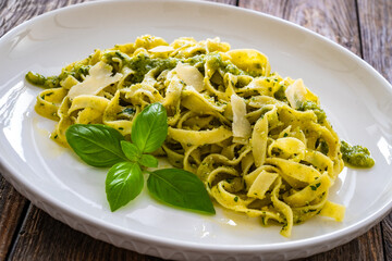 Wall Mural - Pappardelle with basil pesto sauce sprinkled with grated Parmesan cheese served on white plate with spoon and fork on wooden table
