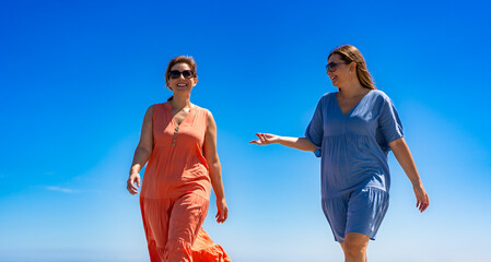 Wall Mural - summer vacation. two pretty middle aged women wearing flowing dresses and sunglasses talking and walking against the blue sky on a beautiful sunny day