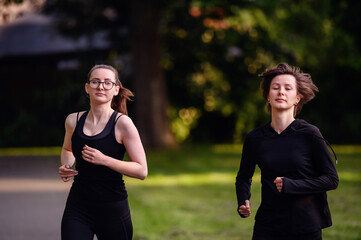 Two young women in black sportswear jogging side by side on a park path, exemplifying fitness, friendship, and a healthy lifestyle.