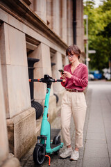 Sticker - Woman in casual attire looking at her phone while standing next to a parked electric scooter on a city sidewalk.