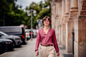 Wall Mural - Woman in casual attire smiling while walking on a city sidewalk lined with parked cars and buildings.