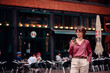 © Иванна Емельянова - Woman in casual attire checking her phone while standing outside a busy cafe with people seated at tables.