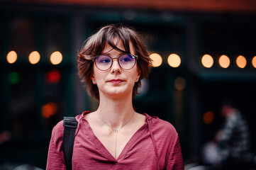 Wall Mural - Close-up of a woman wearing glasses, standing in front of a blurred urban background with lights.