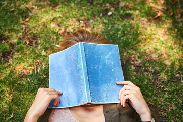 Girl, garden and cover face with book on lawn for reading, fantasy and romance novel or love poetry. Woman, outdoor and sleeping of hobby for romantic literature, knowledge and calm on grass.