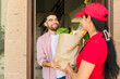 © AntonioDiaz - Cheerful female delivery person in a red uniform handing a grocery bag to a young man at his doorstep