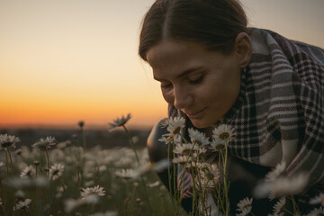 Wall Mural - A woman is kneeling in a field of flowers, smelling them. The sky is orange and the flowers are white.