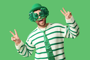 happy young man in leprechaun's hat with party glasses showing victory gesture on green background. 