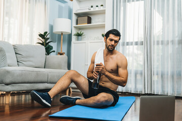 Wall Mural - Athletic and sporty man sitting, resting on fitness mat after finishing home body workout exercise session for fit physique and healthy sport lifestyle at home. Gaiety home exercise workout training.