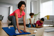 © NDABCREATIVITY - Young woman, mother exercising at home in living room, father playing with kid in background.