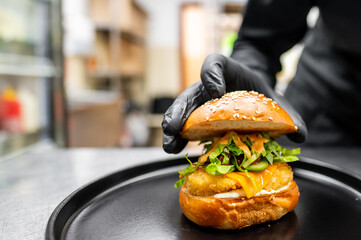 A hand in a black glove holds the top bun of a burger over a plate, showcasing fresh ingredients and appealing presentation