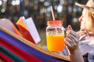 Woman drinking orange smoothie while resting and reading book in hammock. Relax and joy of calm moment in nature