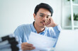 © KANGWANS - A man is sitting at a desk with a laptop and a stack of papers. He is looking at a piece of paper and he is in a state of deep thought