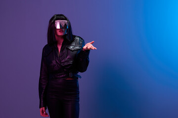 Wall Mural - Confused pretty awesome brunet woman in leather jacket trendy specular sunglasses point hand look aside posing isolated in blue violet color light background. Neon party Cyberpunk concept. Copy space