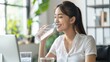 © Vitalii Shkurko - Smiling asian woman drinking water from a bottle while working at a desk with a laptop, surrounded by indoor plants.
