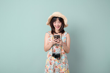 Wall Mural - Travel and vacation concept. Happy Asian woman wearing casual dress and hat with camera while using mobile phone on vacation or travel theme isolated on pastel green background.