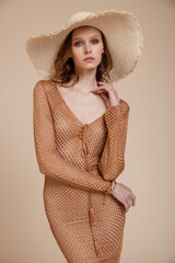 High fashion photo of a beautiful skinny young woman in pretty brown mesh beach dress, hat posing on beige background. Slim figure. Summer style