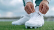 © Iana - A close-up shot of a player's hands tying the laces on their football cleats, with blades of grass and the edge of the pitch visible in the background