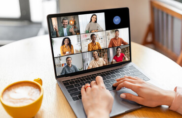Poster - Lady sits at a table in a coffee shop, using a laptop to participate in a video conference with a group of people. The screen shows six video tiles of the participants