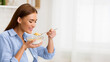 © Prostock-studio - A woman in a blue striped shirt eats cereal from a bowl in her kitchen. She is holding a spoon with a mouthful of cereal and looks happy.