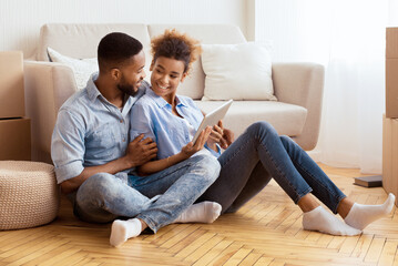 Wall Mural - Young Black Couple Using Digital Tablet Sitting On Floor Among Moving Boxes Relaxing In New Apartment. House Ownership