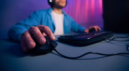 Leinwandbilder - A close-up view of a gamers hand gripping a wired computer mouse, with the keyboard and a blurred figure in the background. The scene is lit by purple and pink lighting, cropped