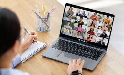Poster - Lady woman sits at a desk, taking notes with a pen and notepad as she participates in a virtual meeting on her laptop. The screen displays a grid of video calls