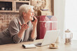 © Prostock-studio - Elderly woman with short, gray hair sits at a kitchen table, talking on her phone. She is smiling and gesturing with her free hand. A laptop is open in front of her and a notebook is lying next to it.