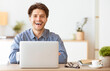 © Prostock-studio - A man sits at his desk with a laptop, smiling brightly and looking at the camera.