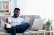 © Prostock-studio - African American man is relaxing on a couch in a living room. He is holding a tablet in one hand and a cup of coffee in the other. He is smiling and looks relaxed.