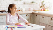 © Prostock-studio - A young girl with long dark hair sits at a white table in a kitchen. She is smiling and looking to her right. There are colored pencils scattered on the table, and a few are in focus, copy space
