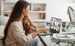 © Prostock-studio - A woman is sitting in her home office, attending a virtual meeting on her laptop. The laptop screen displays a grid of video calls from multiple participants.